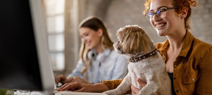 Smiling woman with dog in lap at a computer with woman in the background